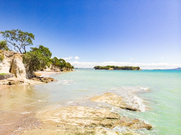 A serene coastal landscape features clear turquoise waters gently lapping against the rocky shoreline, with a distant view of a tree-lined island under a vibrant blue sky. The rocky formations and sandy beach are accentuated by patches of greenery, indicating a natural and untouched environment.