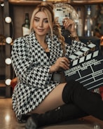 A woman sits confidently in a chic, houndstooth-patterned coat with black knee-high boots, holding a film clapperboard. The background features a disco ball and bottles, with soft lighting creating a glamorous ambiance.