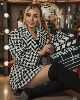 A woman sits confidently in a chic, houndstooth-patterned coat with black knee-high boots, holding a film clapperboard. The background features a disco ball and bottles, with soft lighting creating a glamorous ambiance.