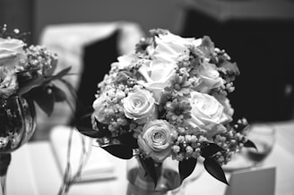 A monochrome photograph of an elegant floral arrangement, primarily consisting of roses and daisies, set in a vase. The background features blurred elements that suggest an indoor setting.