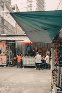 In a narrow alleyway, three people are seated on stools around a table, seemingly engaged in conversation or enjoying a meal. The area is covered by a tarp, and behind them is a small food stall. The scene captures an informal, everyday moment in an urban setting.