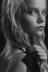 A black and white portrait of a young girl with long hair, gazing directly at the camera. The lighting creates a soft, ethereal atmosphere, highlighting her expressive eyes and delicate features. Her hand is gently holding the strap of her clothing.