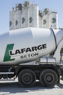 A cement mixer truck with 'Lafarge Beton' branding is parked in front of several large industrial silos. The silos are white with a green logo resembling a stylized lion on them. The scene is brightly lit, suggesting a sunny day.