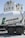 A cement mixer truck with 'Lafarge Beton' branding is parked in front of several large industrial silos. The silos are white with a green logo resembling a stylized lion on them. The scene is brightly lit, suggesting a sunny day.