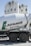 A cement mixer truck with 'Lafarge Beton' branding is parked in front of several large industrial silos. The silos are white with a green logo resembling a stylized lion on them. The scene is brightly lit, suggesting a sunny day.