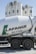 A cement mixer truck with 'Lafarge Beton' branding is parked in front of several large industrial silos. The silos are white with a green logo resembling a stylized lion on them. The scene is brightly lit, suggesting a sunny day.