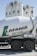 A cement mixer truck with 'Lafarge Beton' branding is parked in front of several large industrial silos. The silos are white with a green logo resembling a stylized lion on them. The scene is brightly lit, suggesting a sunny day.