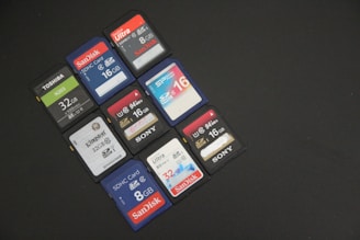 A group of SD cards from various brands like SanDisk, Toshiba, Kingston, and Sony are arranged on a black surface. Each card displays its storage capacity, with sizes ranging from 8GB to 32GB. The colors of the cards vary, including black, blue, red, and white.