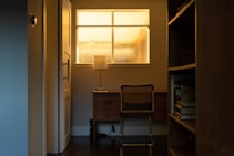 A warmly lit room features a wooden desk with a table lamp illuminating the space with a soft glow. A single chair with a woven backrest sits pushed under the desk. A window with textured glass allows ambient light to filter through, creating a cozy atmosphere. Books and other items are visible on a nearby shelf.