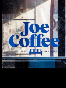 A glass window with bold blue text that reads 'Joe Coffee,' alongside a small graphic of a bench. The words 'EST. 2003' and 'NEW YORK' are printed beneath.