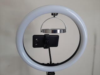 A circular LED ring light is mounted on a stand, with a smartphone attached in the center of the ring using a holder. The setup appears to be prepared for recording or streaming purposes.
