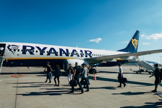 Passengers are walking towards a Ryanair airplane on the tarmac, preparing to board via a stairway connected to the aircraft's front entrance. The sky is clear with a few clouds, and the overall atmosphere is sunny and bright.