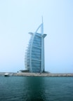 A modern, sail-shaped luxury hotel rises prominently against a clear blue sky. Situated on an artificial island, it is connected to the mainland by a bridge. Palm trees line the base of the structure, with a small yacht docked near the shore. The building features a distinctive architectural design with a sleek, curved facade.