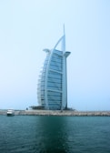 A modern, sail-shaped luxury hotel rises prominently against a clear blue sky. Situated on an artificial island, it is connected to the mainland by a bridge. Palm trees line the base of the structure, with a small yacht docked near the shore. The building features a distinctive architectural design with a sleek, curved facade.