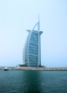 A modern, sail-shaped luxury hotel rises prominently against a clear blue sky. Situated on an artificial island, it is connected to the mainland by a bridge. Palm trees line the base of the structure, with a small yacht docked near the shore. The building features a distinctive architectural design with a sleek, curved facade.