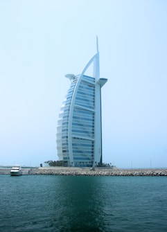 A modern, sail-shaped luxury hotel rises prominently against a clear blue sky. Situated on an artificial island, it is connected to the mainland by a bridge. Palm trees line the base of the structure, with a small yacht docked near the shore. The building features a distinctive architectural design with a sleek, curved facade.