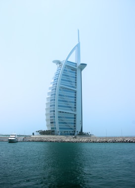 A modern, sail-shaped luxury hotel rises prominently against a clear blue sky. Situated on an artificial island, it is connected to the mainland by a bridge. Palm trees line the base of the structure, with a small yacht docked near the shore. The building features a distinctive architectural design with a sleek, curved facade.