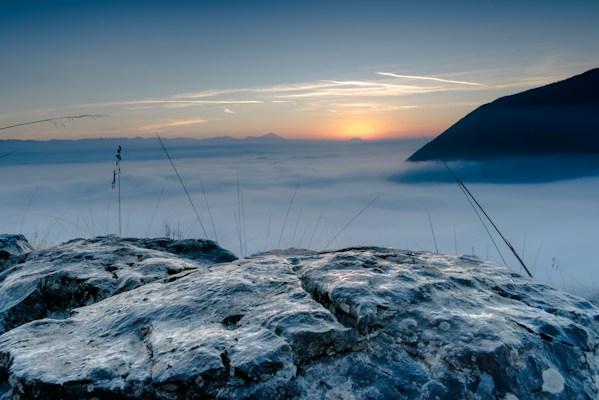 A misty sunrise over a mountainous landscape, with the sun partially visible on the horizon creating a soft glow. The foreground is dominated by rocky terrain with some blades of grass protruding. The expansive view includes a thick layer of fog covering the valley below, creating a serene and tranquil ambiance.