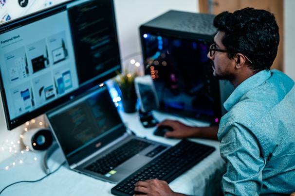A person wearing glasses is working on a computer setup with multiple screens. The large monitor displays web designs, and the laptop screen shows code. The workspace is lit with soft ambient lighting, and a gaming desktop is visible beside the person.