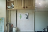 A kitchen with wooden cabinets above a tiled backsplash. A hand-drawn picture is taped to the tiles, showing a leaf and a profile of a face. Below, there are various items including a potted plant, some books, and a pot with a lid on the stove.