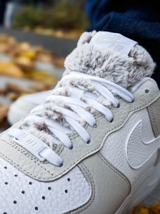 A close-up of a sneaker featuring textured leather and fur-like material around the ankle. The laces are white, and a logo is visible near the top. The sneaker appears to be on a person's foot, with autumn leaves scattered in the background.