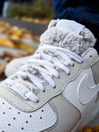 A close-up of a sneaker featuring textured leather and fur-like material around the ankle. The laces are white, and a logo is visible near the top. The sneaker appears to be on a person's foot, with autumn leaves scattered in the background.