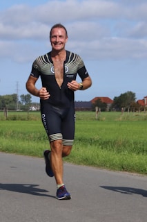 A man is running outdoors on a paved path surrounded by green fields and distant trees. He is wearing a black athletic outfit with a partially unzipped top, featuring a design on the shoulders and legs. There's a smile on his face, suggesting enjoyment or accomplishment.