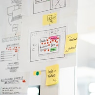 A whiteboard or wall is filled with paper sheets and sticky notes containing diagrams, sketches, and notes. Prominent features include hand-drawn flowcharts with symbols and arrows, as well as several colored sticky notes with handwritten words like 'Feedback' and 'Infos + Features'. The central sheet displays a layout with text and colored dots, possibly representing an interface design or process.
