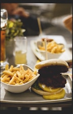 A plate of crispy french fries served alongside a large burger stacked with lettuce and mustard, placed on a table outside. Beverages in clear glasses are in the background.