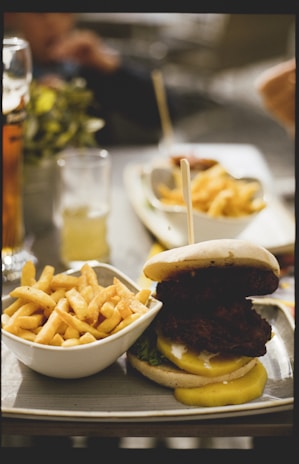 A plate of crispy french fries served alongside a large burger stacked with lettuce and mustard, placed on a table outside. Beverages in clear glasses are in the background.