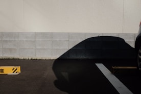 A parking lot scene with a concrete wall in the background and the shadow of a car prominently cast on the gray tiles. A yellow parking stopper with black stripes is positioned on the asphalt, and part of a car is visible on the right side.