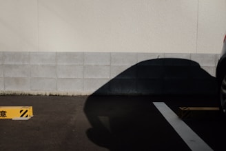 A parking lot scene with a concrete wall in the background and the shadow of a car prominently cast on the gray tiles. A yellow parking stopper with black stripes is positioned on the asphalt, and part of a car is visible on the right side.