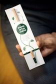 A hand is holding a rectangular package that contains a pencil. The packaging features botanical illustrations and text in Italian. The pencil appears to have a green capsule at one end, possibly indicating a plantable feature.