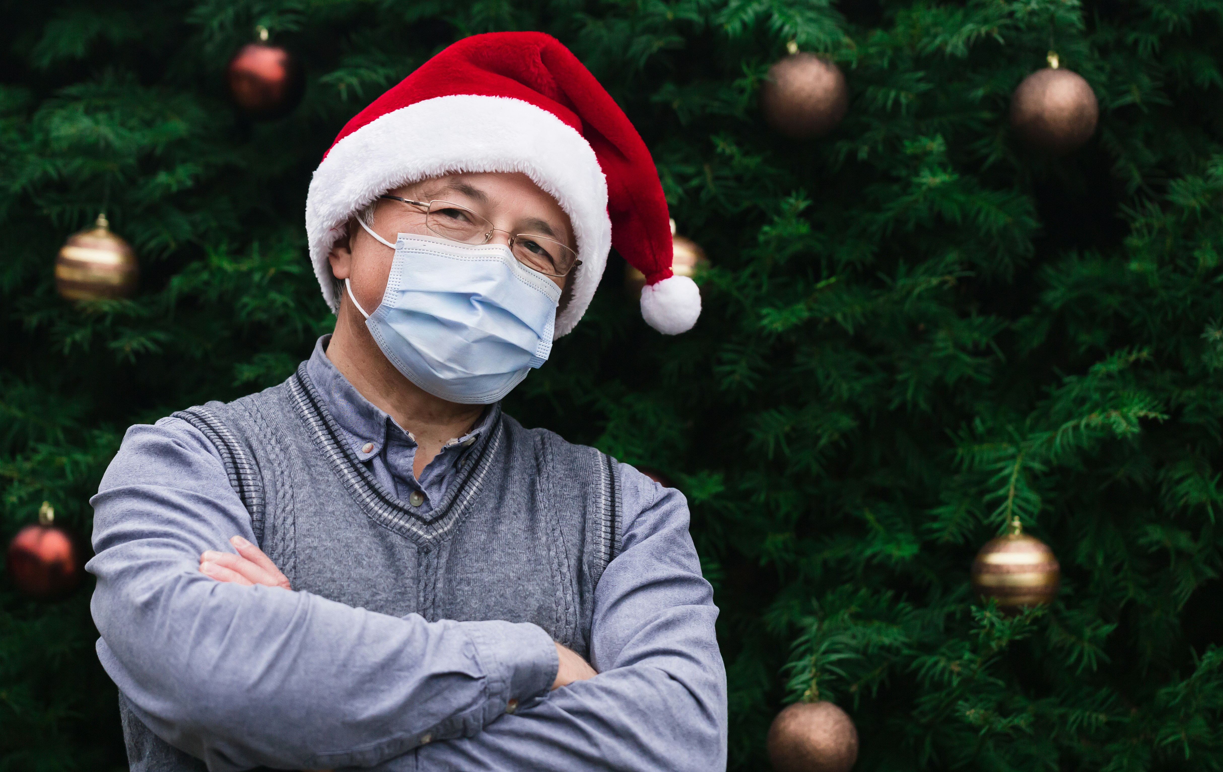 Corona Christmas. Close up Portrait of senior man wearing a santa claus hat and medical mask with emotion. Against the background of a Christmas tree. Coronavirus pandemic