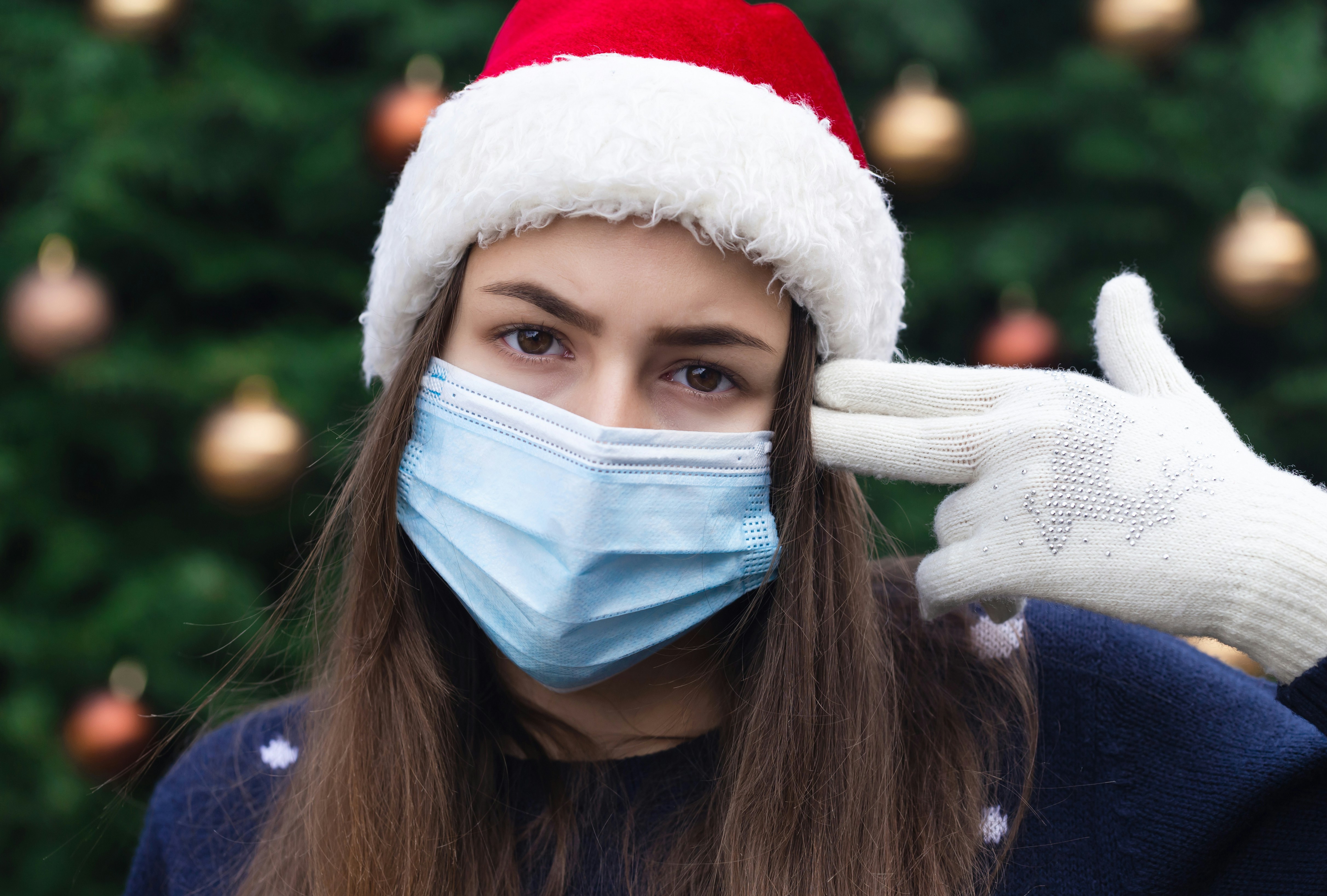 Close up Portrait of woman wearing a santa claus hat and medical mask with emotion. Against the background of a Christmas tree. Coronavirus pandemic