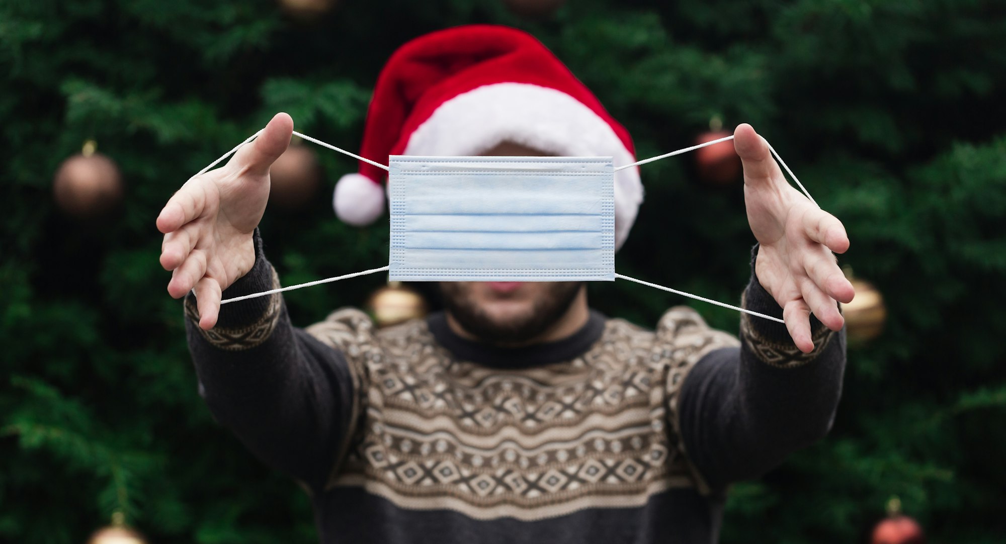 Anonymous Christmas. Faceless Portrait of man wearing a santa claus hat, xmas sweater and hold medical mask with emotion. Against the background of a Christmas tree. Coronavirus pandemic