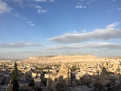 A panoramic view of a town nestled in a valley with unique rock formations known as fairy chimneys. The landscape is mostly arid with sporadic greenery, and the town's architecture is characterized by low-rise buildings. The sky is partly cloudy, enhancing the picturesque setting with its expansive blue hues.