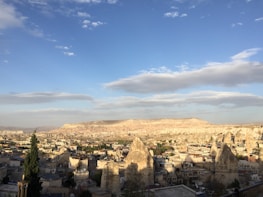A panoramic view of a town nestled in a valley with unique rock formations known as fairy chimneys. The landscape is mostly arid with sporadic greenery, and the town's architecture is characterized by low-rise buildings. The sky is partly cloudy, enhancing the picturesque setting with its expansive blue hues.