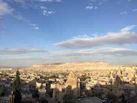 A panoramic view of a town nestled in a valley with unique rock formations known as fairy chimneys. The landscape is mostly arid with sporadic greenery, and the town's architecture is characterized by low-rise buildings. The sky is partly cloudy, enhancing the picturesque setting with its expansive blue hues.
