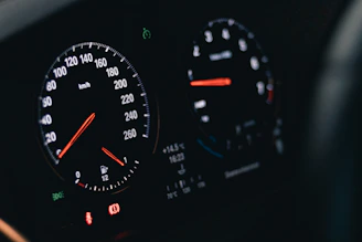 A close-up of an automobile dashboard displaying two circular gauges with illuminated dials and needles. The left gauge is a speedometer marked in kilometers per hour, and the right gauge includes a tachometer and fuel indicator. Various indicator lights are visible at the bottom of the display.