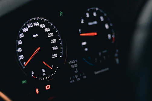 A close-up of an automobile dashboard displaying two circular gauges with illuminated dials and needles. The left gauge is a speedometer marked in kilometers per hour, and the right gauge includes a tachometer and fuel indicator. Various indicator lights are visible at the bottom of the display.