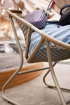 A person is seated on a woven chair, with a colorful patterned handbag resting on their lap. The chair features a sturdy, light beige frame with intricate woven details, providing a contrast against the person's light-colored clothing. The background includes blurred, warm tones suggestive of an indoor setting.