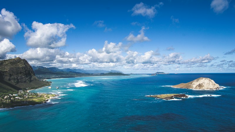 Coastal headland with offshore islets and reef seen from a high clifftop, deep blue water under scattered clouds