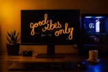 A dimly lit workspace with a monitor displaying the text 'good vibes only' in a warm, glowing script. To the left, a small potted plant adds a touch of greenery. On the desk, a camera and a mug are visible, suggesting hobbies or interests. The computer setup includes a tower with blue LED lights visible on the right, adding a modern, tech-savvy vibe.