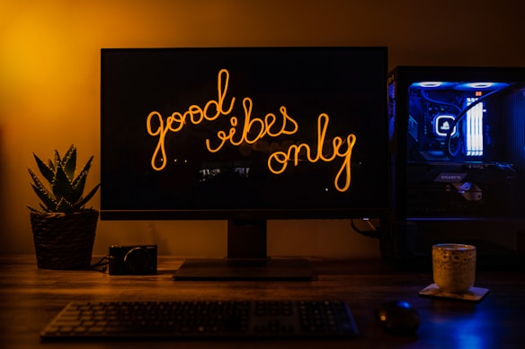 A dimly lit workspace with a monitor displaying the text 'good vibes only' in a warm, glowing script. To the left, a small potted plant adds a touch of greenery. On the desk, a camera and a mug are visible, suggesting hobbies or interests. The computer setup includes a tower with blue LED lights visible on the right, adding a modern, tech-savvy vibe.
