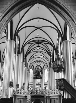 A grand and imposing interior of a cathedral, showcasing high vaulted arches, tall columns, and ornate decorations. The ceiling features intricate ribbed vaulting, while elaborate chandeliers hang from above. The architecture is reminiscent of Gothic style, with detailed ornamentations and a feeling of vastness. The image is in black and white, highlighting the contrasts and structural details.