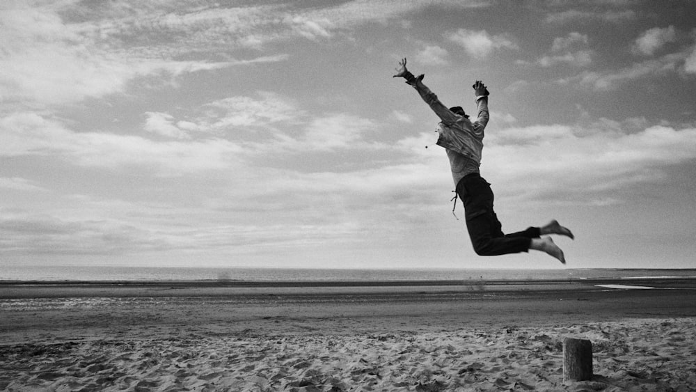 foto in scala di grigi di un uomo che salta sulla spiaggia
