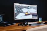 A modern workspace features a large curved monitor displaying a web agency page, surrounded by various items including a laptop, a smartphone, a wireless keyboard, headphones, and a stylus pad. The setup is neatly arranged on a wooden desk against a dark wall, creating a sleek and professional look.