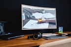 A modern workspace features a large curved monitor displaying a web agency page, surrounded by various items including a laptop, a smartphone, a wireless keyboard, headphones, and a stylus pad. The setup is neatly arranged on a wooden desk against a dark wall, creating a sleek and professional look.