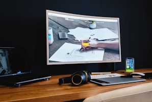 A modern workspace features a large curved monitor displaying a web agency page, surrounded by various items including a laptop, a smartphone, a wireless keyboard, headphones, and a stylus pad. The setup is neatly arranged on a wooden desk against a dark wall, creating a sleek and professional look.